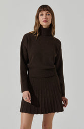 A woman is wearing a dark brown, chunky knit turtleneck sweater with long sleeves and a ribbed hem. She pairs it with a matching dark brown ribbed knit pleated skirt.